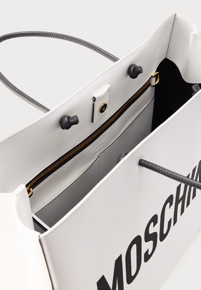 Sac cabas blanc Moschino avec poignées noires, poche intérieure zippée avec fermeture éclair dorée et fermeture à pression magnétique.