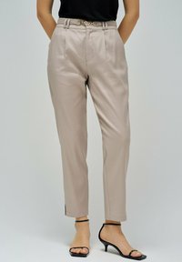 Mujer vistiendo pantalones de talle alto en color beige con un cinturón delgado, top sin mangas negro y sandalias de tacón negro con tiras, de pie contra un fondo gris.