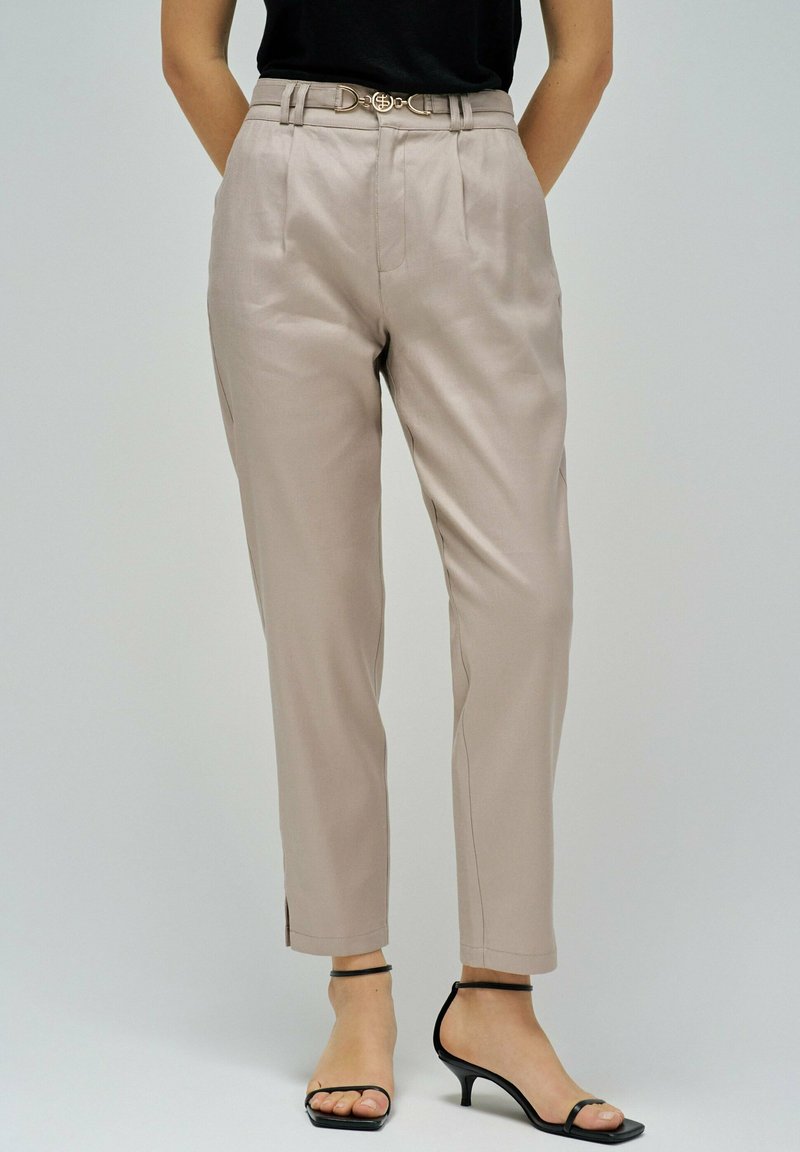 Mujer vistiendo pantalones de talle alto en color beige con un cinturón delgado, top sin mangas negro y sandalias de tacón negro con tiras, de pie contra un fondo gris.
