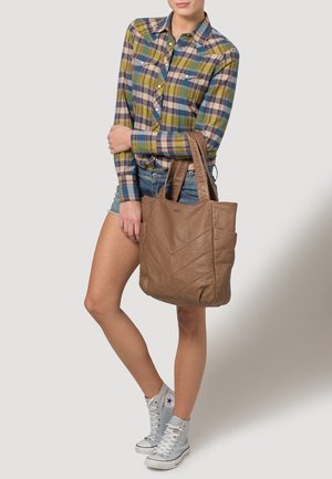 Sac en cuir marron avec motif en chevrons, poignées solides et finition texturée. Le modèle porte une chemise à carreaux et un short en denim.