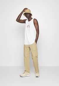 Top blanco sin mangas con texto rojo, pantalones cargo beige y un sombrero tipo cubo beige. Zapatillas beige completan el atuendo. Estilo streetwear casual.
