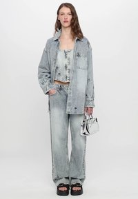 Šviesiai mėlynas džinsinis švarkas ir kelnių komplektas su kryžiaus siuvinėjimu. Suderintas trumpas topas, blizgus sidabro spalvos rankinukas ir juodi aukšti sandalai.