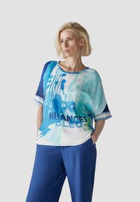 Kurze Ärmeloberteil mit einem blau-türkisen Grafikdruck, der den Text "NUANCES BLEU" zeigt, und einer lockeren Passform. Kombiniert mit blauen Hosen.