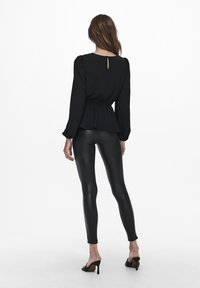 Top nero a maniche lunghe con vita a peplo e dettaglio a chiave sul retro, abbinato a leggings neri lucidi e sandali con punta aperta.