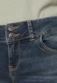 Jeans aus Denim in dunkler blauer Farbe mit goldenen Nähten. Zwei Knöpfe am Bund und eine seitliche Tasche sichtbar. Glatte Textur.