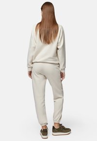 Jasno beżowa bluza i pasujące dresy z elastycznymi mankietami; tkanina wydaje się być miękka. Brązowe sneakersy z teksturowanymi akcentami i białymi podeszwami.