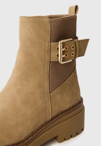 Botte de cheville beige avec un panneau élastique texturé marron, détails de boucle en métal doré et une semelle épaisse à rainures. Matériau en simili cuir lisse.