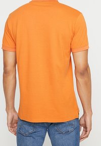 Polo shirt in cotone arancione con colletto classico, maniche corte e spacco laterale per facilita' di movimento. Dettagli di cucitura visibili.