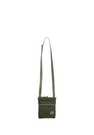 Kleine olijfgroene crossbodytas met verstelbare riem en voorvak met ritssluiting, met een rechthoekig zwart-wit merketiket.