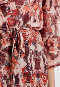 Vestido fluido con un patrón de paisley floral en rojo y azul marino sobre un fondo beige claro, con una cinta de tela en la cintura y una tela texturizada.