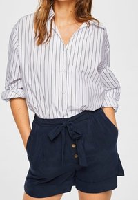Bílá košile na knoflíky s modrými vertikálními pruhy; spárovaná s námořnickými šortkami s vysokým pasem, které mají zavazovací pásek a přední knoflíky.