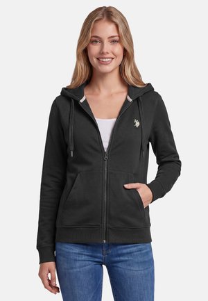 Smiling woman wearing a black zip-up hoodie with front pockets and a white undershirt, paired with blue jeans, standing against a plain background.