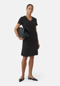 Black short-sleeve dress with a V-neck, smooth texture, knee-length. Model holds a black leather handbag; wearing black pointed flats with slingback.