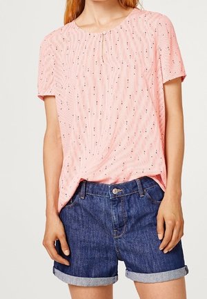 Persona che indossa una camicetta rosa a maniche corte con piccoli pois e pantaloncini di jeans blu risvoltati, in piedi con le mani rilassate lungo i fianchi.