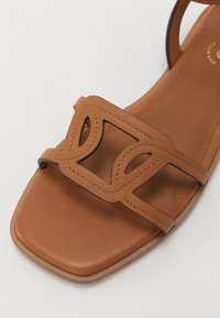 Sandale plate en cuir marron avec un bout carré et une bride géométrique découpée sur le dessus du pied.