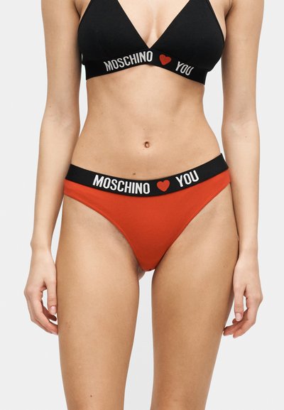 Γυναίκα που φοράει μαύρο τριγωνικό σουτιέν και κόκκινο εσώρουχο με μαύρες ζώνες στη μέση με το κείμενο "MOSCHINO" και κόκκινο σύμβολο καρδιάς.