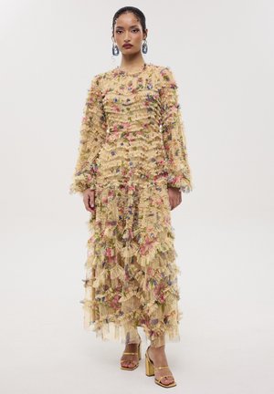 Mujer con un vestido largo beige con volantes florales en capas, de pie con las manos relajadas y con sandalias doradas de tacón alto.