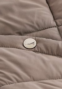 Tissu matelassé beige avec un petit bouton rond en argent gravé d'un logo montagne et d'un texte sur une surface matelassée.
