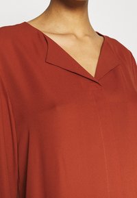 Blouse couleur rouille avec un col en V, présentant un tissu subtilement texturé et un détail de couture simple à l'avant.