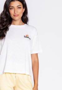 Bílé bavlněné tričko s volným střihem, které má vyšívané logo "Paradise" a design duhy na levé straně hrudi.