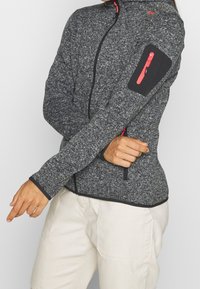 Veste gris zippée en matériau texturé avec des accents noirs. Présente une petite poche zippée sur le bras et des détails de fermeture éclair rouges.