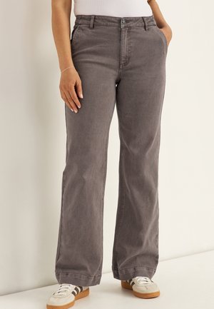 Person trägt hochgeschnittene, weit geschnittene graue Hosen, weiße Turnschuhe mit braunen Sohlen und ein weißes Oberteil, steht mit einer Hand in der Tasche.