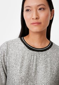 Top in paillettes argentate con superficie testurizzata. Presenta una scollatura a costine nera con strisce bianche. Dettaglio della scollatura e della parte superiore del petto.