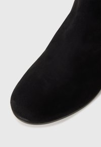 Bottine en daim noir avec bout arrondi et semelle basse sur un fond gris clair.