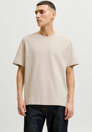 Ung mand med kort hår iført en ensfarvet beige t-shirt og mørke jeans, står foran en lys grå baggrund og kigger mod venstre.
