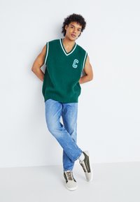 Pulover fără mâneci, verde cu dungi, cu decolteu în V și margine albă, având un litera "C" pe piept; asortat cu blugi decolorați și pantofi sport.