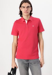 Polo en coton rouge avec un col blanc contrastant, manches courtes et un petit logo sur la poitrine. Porté avec un pantalon en denim gris.