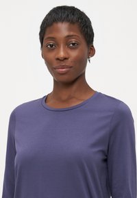 Självsäker kvinna med kort hår som bär en lila långärmad skjorta och små hoopörhängen, mot en enfärgad ljus bakgrund.
