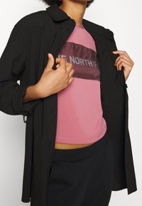 Camisa negra de botones sobre una camiseta gráfica rosa con un diseño de malla, con pantalones negros visibles a la altura de la cintura. No se especifica el tono de piel.