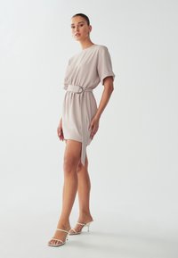 Korte beige jurk met losse korte mouwen, cinched taille met een zilveren ringriem. Het model draagt het met open teen witte hakken.