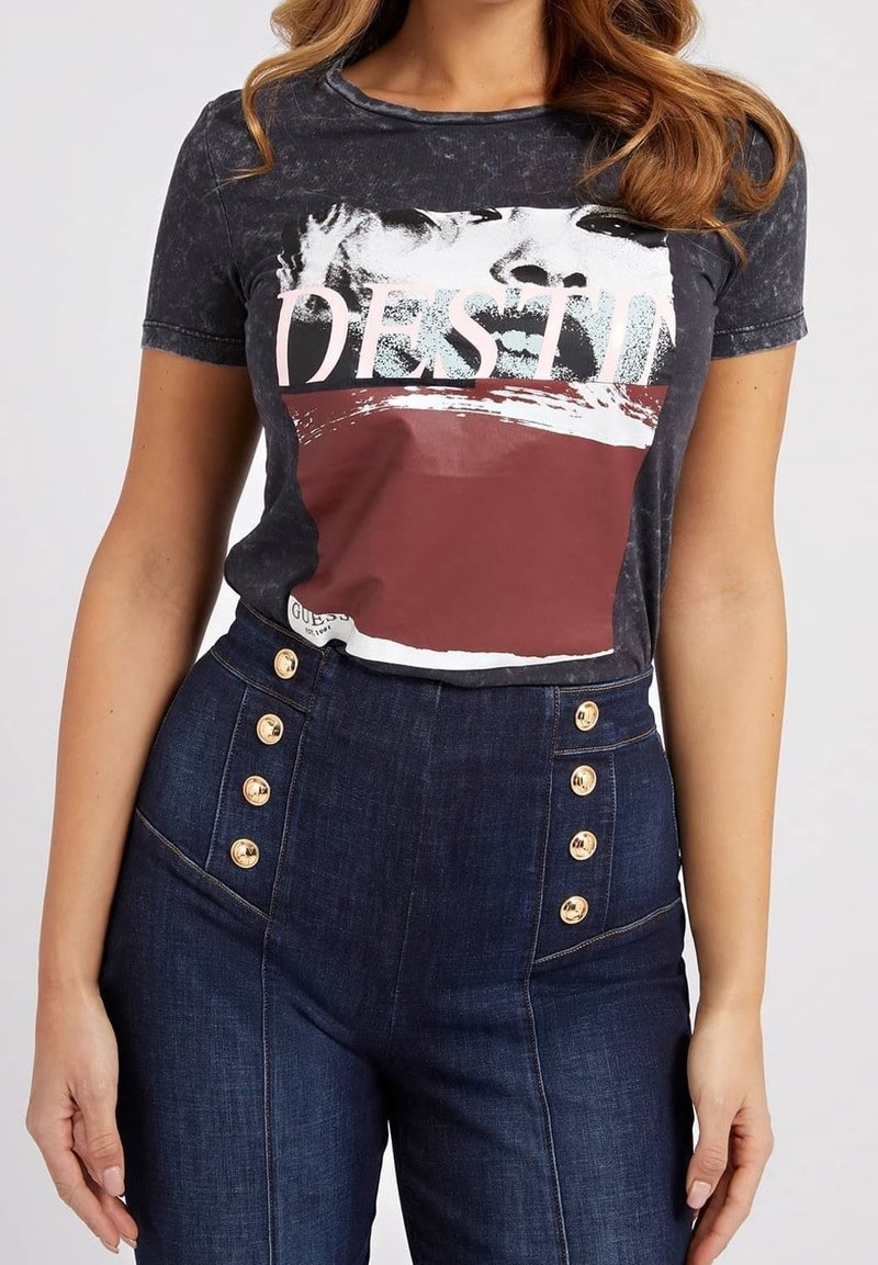 Femme portant un t-shirt graphique noir avec le texte "DESTIN" et un motif visage, associé à un jean bleu foncé taille haute avec des boutons dorés.