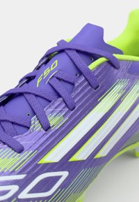 Crampon de football violet avec une tige texturée, des accents verts et des rayures blanches. Les lacets sont visibles sur le dessus, avec un logo "F50".