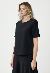 Kobieta o blond włosach, ubrana w gładką czarną bluzkę z krótkim rękawem i dopasowaną czarną spódnicę, stojąca na białym tle, zwrócona w lewo.