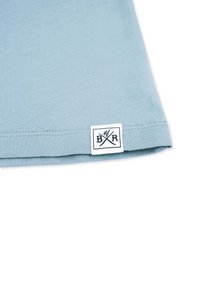 Lyseblå bomuld t-shirt med en glat tekstur, der har et firkantet mærke med "BR" og krydsede redskaber i marineblå ved kanten.