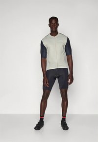 Maillot de cyclisme ajusté gris clair avec manches courtes bleu marine et fermeture éclair frontale. Associé à un short noir et des chaussures de cyclisme noires.