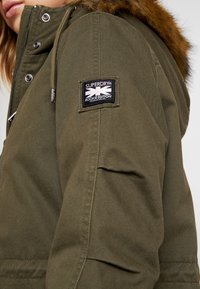 Oliwkowy parka z futrzanym wykończeniem na kapturze, z logo na rękawie i zamkiem przy frontowym zapięciu.