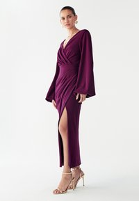 Lange pruimkleurige jurk met een diepe V-hals, gedraaide lijfje, vloeiende klokvormige mouwen en een hoge split, gecombineerd met gouden sandalen met bandjes. Gladde stoftextuur.