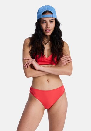 Costum de baie roșu, compus dintr-un top cu decolteu rotund și chiloți cu talie înaltă. Prezintă un material neted și un design mulat. Purtați cu o șapcă albastră.