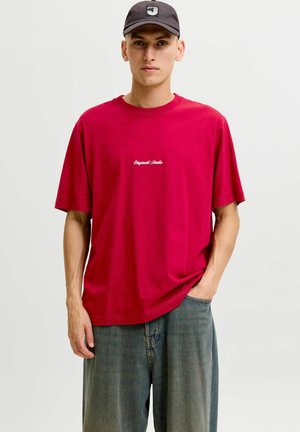 Ung mand iført en løs rød t-shirt med teksten "Original Studio", løstsiddende blå jeans og en sort kasket med et hvidt bogstav J.