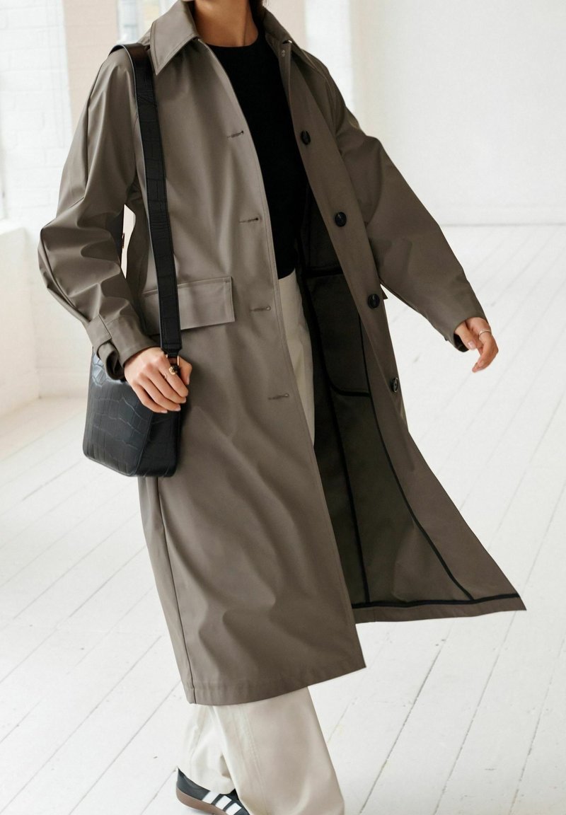Personne portant un long manteau gris, un haut noir, un pantalon blanc et un sac bandoulière noir, debout dans un espace intérieur lumineux et blanc.
