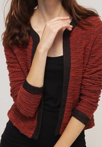 Rød tekstureret cardigan med mørk kant, der har en afslappet pasform og ærmer til albuen. Materialet ser blødt ud med et subtilt bølgemønster.