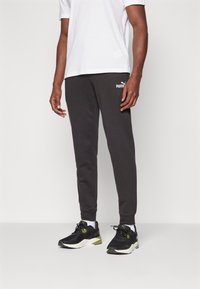 Black fleece joggers with an elastic waistband, ribbed cuffs, and a small white Puma logo on the left thigh. Paired with black athletic sneakers.