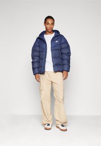 Casaco puffer azul-marinho com capuz, apresentando costura horizontal e um logotipo, combinado com calças cargo bege e ténis brancos com detalhes azuis.