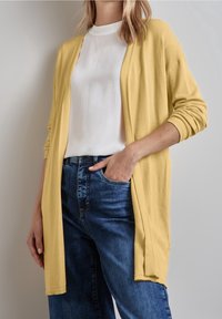 Žlutý pletený cardigan s otevřeným předním dílem, dlouhými rukávy a žebrovanými manžetami, nošený přes bílý top a modré džíny. Měkká textura, ležérní vzhled.