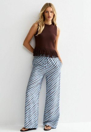 Młoda kobieta w brązowej kamizelce bez rękawów z frędzlami, szerokich jasnoniebieskich spodniach z ukośnymi paskami oraz czarnych sandałach, stojąca na tle białej ściany.