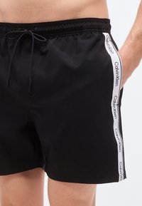 Μαύρα μαγιό σορτς με ελαστική μέση και κορδόνι, καθώς και λευκή ρίγα με λογότυπο στα πλαϊνά. Κατασκευασμένα από ελαφρύ, απαλό ύφασμα.